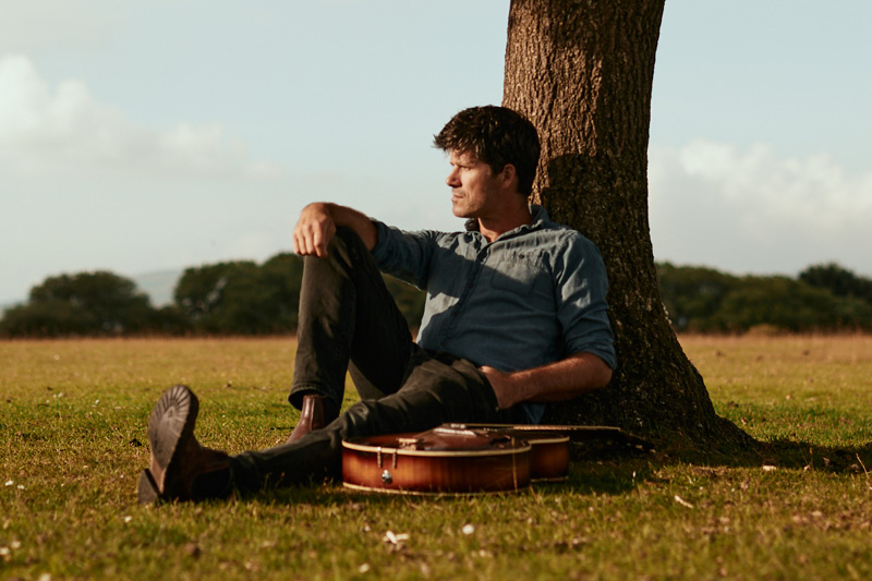 Seth Lakeman sitting on the grass with his back leaning up against a tree