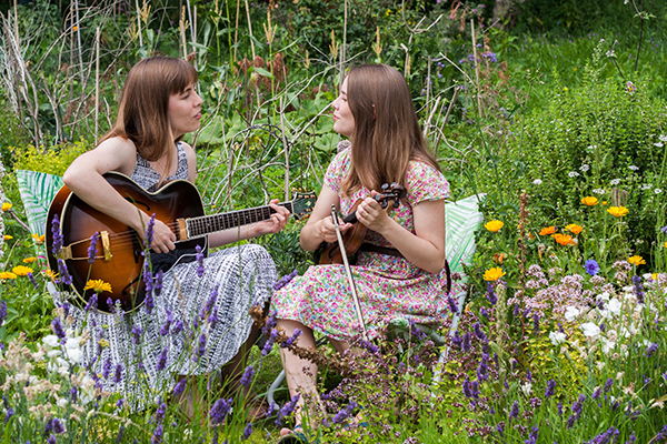 The Carrivick Sisters sat in amongst grass and flowers, facing each other singing and playing their instruments 