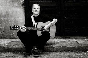 Black and white image of Jason Hinchey sitting on a concrete step, playing his guitar