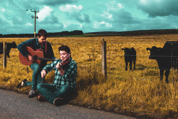 An image of Arthur & Kerran. Arthur is sat down playing violin and Kerran is knelt down next to him playing guitar. They are on the side of a road with a big field behind them. There are 2 cows in the frame. The image is high contrast, the grass is yellow and the sky is blue with clouds in it.