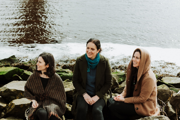 Image of The Henry Girls. 3 women are sat on rocks by the sea edge. They are looking off to the left and the ocean is behind them.