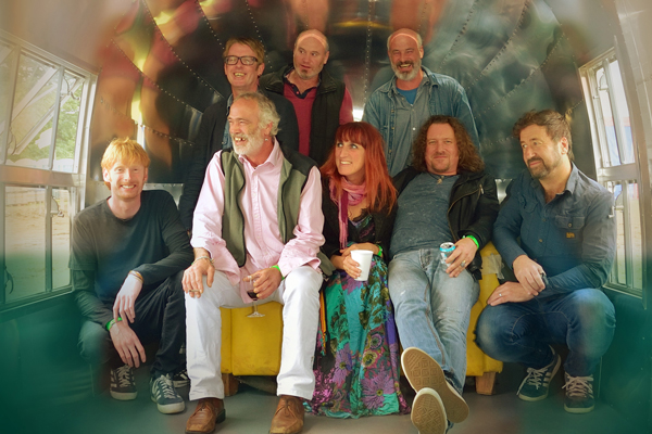 8 members of Kíla are in the image, all with big smiles. They are inside a silver bullet style caravan. 3 of them are sat on a yellow sofa and the rest are either crouched to the side or stood behind the sofa. A few of them have drinks in hand and the silver caravan roof above them is creating a colourful reflection. 