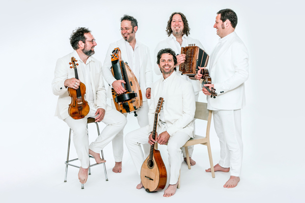 Image of Le Vent du Nord. 5 members of Le Vent du Nord are wearing all white suits and white shirts. They are bare foot and each holding an instrument. Some of them are looking at each other and some are looking at the camera. 1 is sat on a stool, 1 is sat on a chair, and the others are stood.