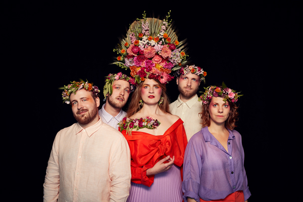 Image of Marvara. There are 5 band members in the image wearing bright coloured clothing and flower crowns. The lead female artist has a larger flower headpiece which stands taller. They all have orange and pink eye makeup on, and are stood up, facing the camera.