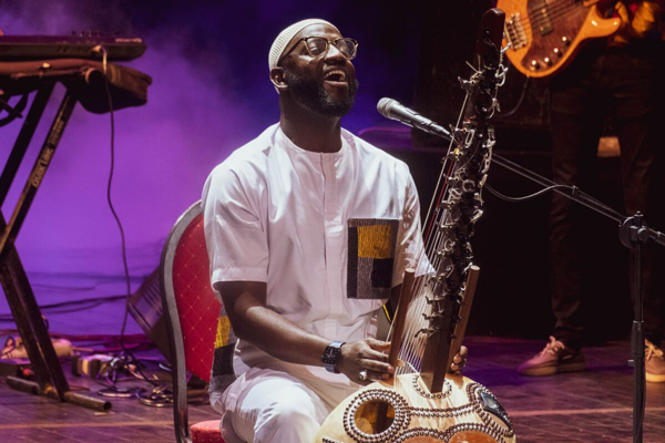 Image for Seckou Keita. Seckou Keita is sitting down on a chair on a stage, singing and playing the kora instrument.