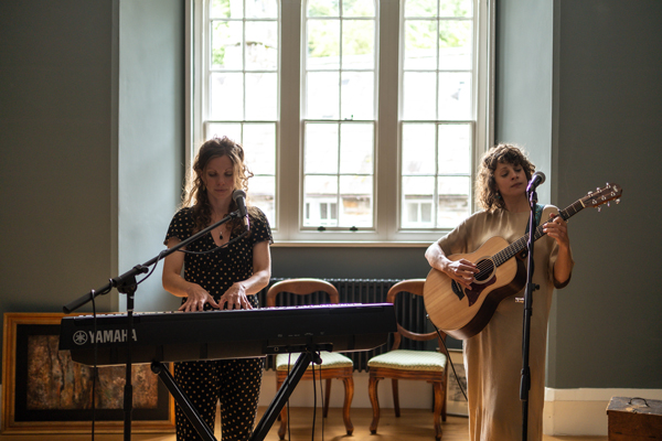 Image of Suthering. The female duo are performing indoors. They are performing in front of a large window. One member is playing piano and the other is playing guitar.