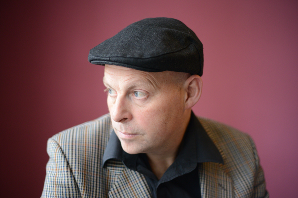 Close up image of Stompin' Dave. He is wearing a black flat cap, a black shirt, and a checkered suit jacket. The background is a pinky colour.