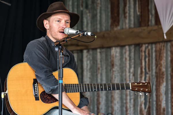 Ben Morgan-Brown (Winner 2019). Performing at Purbeck Valley Folk Festival with hat on, singing into microphone, sat down, with guitar rested on knee.