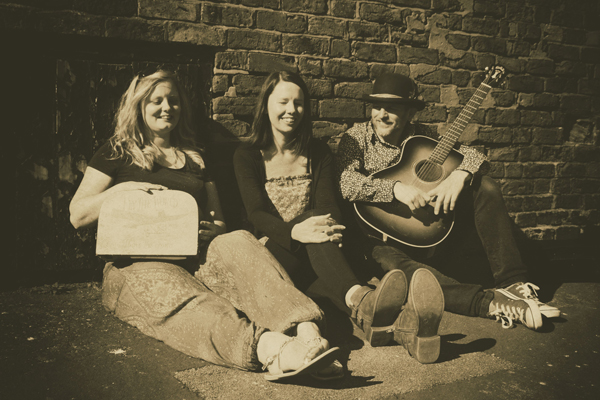 Image of 14 Wolves. The image is in a sepia tone. They are sat on the concrete floor with their backs resting up against a brick wall. There are two females on the left, and a male holding a guitar on the right. 
