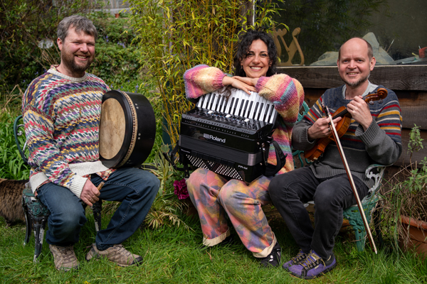 The trio (2 males, 1 female) are sat in a garden with their instruments. 