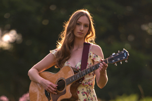 Image of Anna Renae. She is playing her acoustic guitar. The sun is setting and she's stood in a field with lavender coloured flowers. 