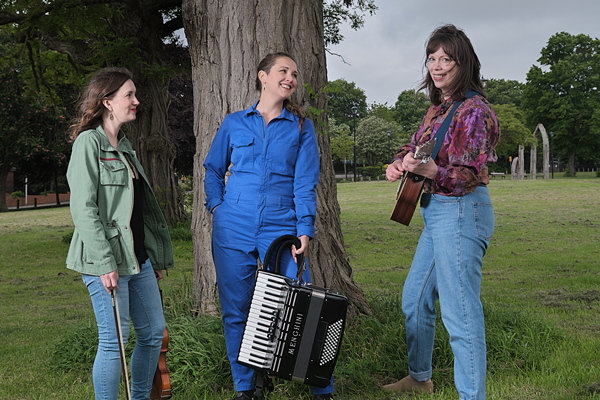 The female trio are stood outside with their instruments, in front of a tree. 