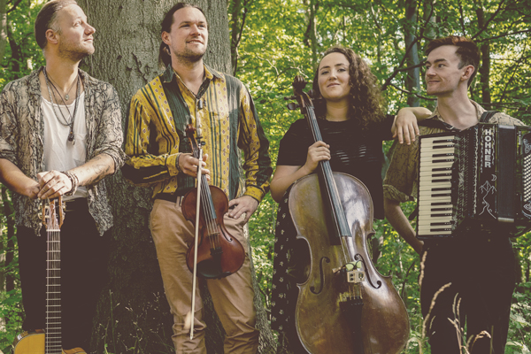 Good Trouble image. 4 band members are stood in a forest holding their instruments. 