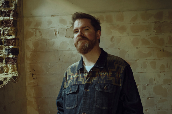 John Smith image. John is staring straight into the camera with a gentle smile on his face. He's wearing a jean jacket, has an auburn beard, and is stood in front of a brick wall. 