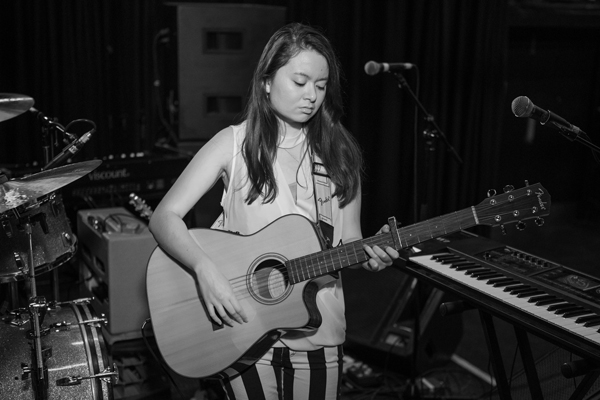 Laura Loh image. A black and white image of Laura holding / playing an acoustic guitar. She is surrounded by other instruments (drum kit, key board, microphones). It looks like she's in a recording studio. 