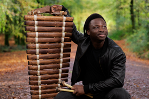 N'famady Kouyate crouched down next to his balafon (traditional wooden xylophone). He is wearing all black and there is a forest in the background of the image.