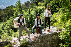 3 male musicans stood on an old brick wall with a mountain forest behind them 