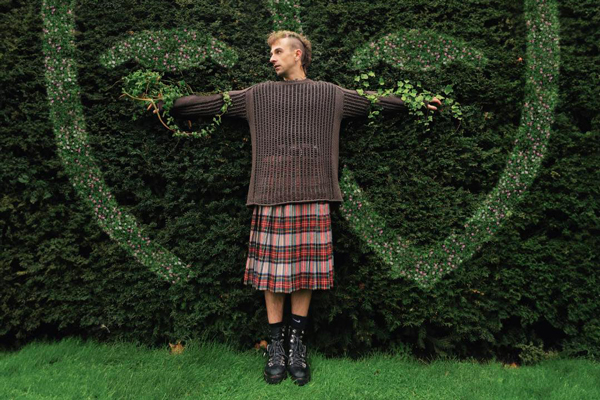 Eli Greeneyes image. Eli is stood on grass in front of a tall green hedge. His arms are stretched out either side of him, with green vines wrapped around his arms. He's looking off to the left. His hair is cut in a mohawk style. He's wearing a brown long-sleeve jumper, a red tartan kilt / skirt, and black boots. 