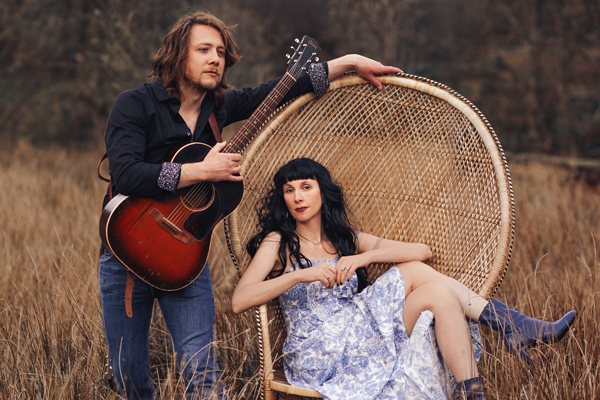 Hannah Sanders & Ben Savage image. The duo are outside in long grass. Hannah is wearing a white and blue dress. She has long black hair with a fringe. She is sat with her legs crossed in a large wicker chair. Ben is stood up next to the chair holding his guitar. He has shoulder length brown hair and is wearing a black shirt and blue jeans.