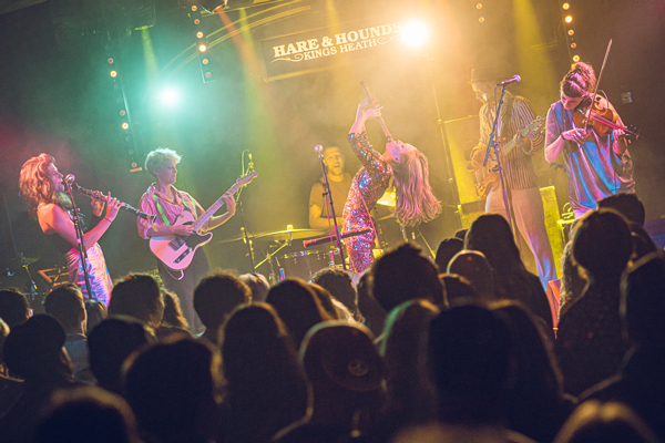 Bonfire Radicals image. </p>
<p>The band are on stage performing. We can see audience members in he foreground. The image is colourful and lively. 