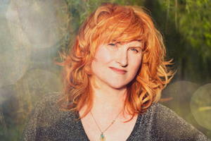 Eddi Reader image. A headshot image of Eddi. The background is green and unfocussed. Eddi has bright orange hair, a soft smile, and blue eyes. Eddi is wear a dark sparkly top, and a gold necklace.