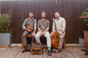 Toby Shaer, Archie Moss & Jack Warnock image. The trio are sat outside in front of a brown garden fence. They are holding their instruments and looking plainly towards the camera.<br />
