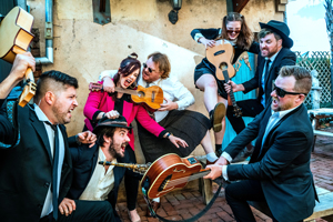 Ukulele Death Squad image. The 7 band members are outside an old building. Most of them are in black and white clothing, like suits and shirts. They are posed playfully, like they are fighting and hitting each other with their stringed instruments.