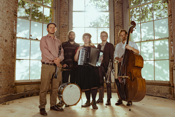 Around About Dusk image. The 5 members are posing with their instruments in front of 3 large floor to ceiling length windows.