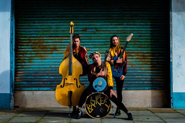 Baskery image. </p>
<p>The trio are posed outside a vibrant blue rusty garage door. They are posing with their instruments.