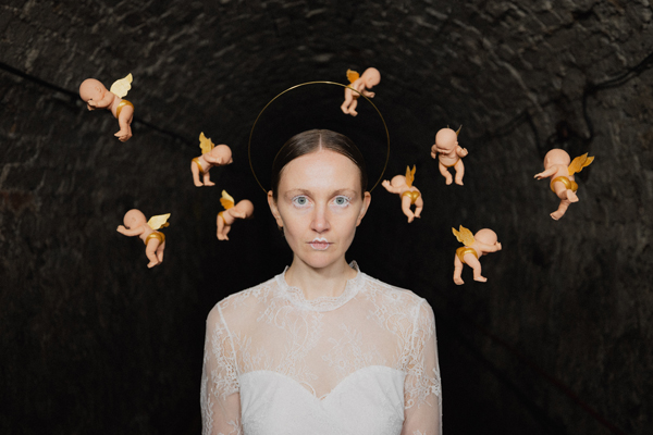 Frankie Archer image. </p>
<p>Frankie is wearing a white dress, white mascara, and white lip liner. The background is dark, and there are little cherubs floating behind her head.
