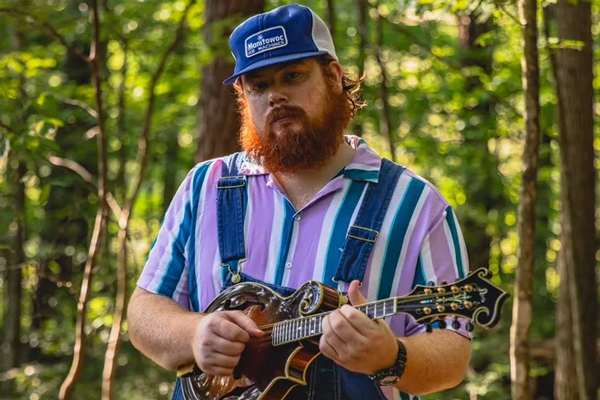 Michael Prewitt image. </p>
<p>Michael is stood in the forest holding his mandolin. He is wearing a blue baseball / trucker style hat, a purple, white, and blue stripy shirt, and blue dungarees. 