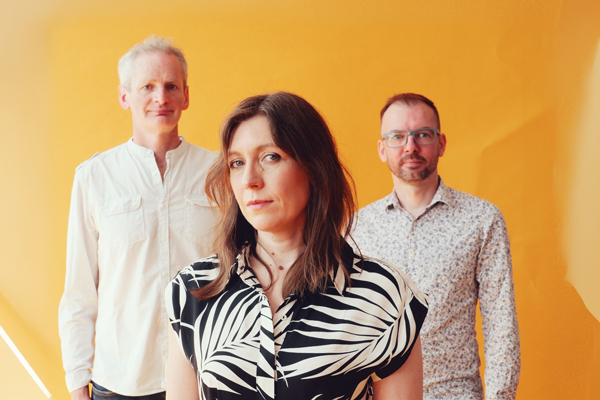 Rachael McShane & The Cartographers image. </p>
<p>The trio have a bright orange backdrop. They are wearing neutral colours (white, black, grey). 
