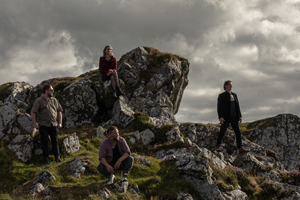 The Roving Crows image. The band are outside on a cloudy day. They are on a cliffside, looking out. The sun is breaking through the grey clouds, giving the image a dramatic feel.