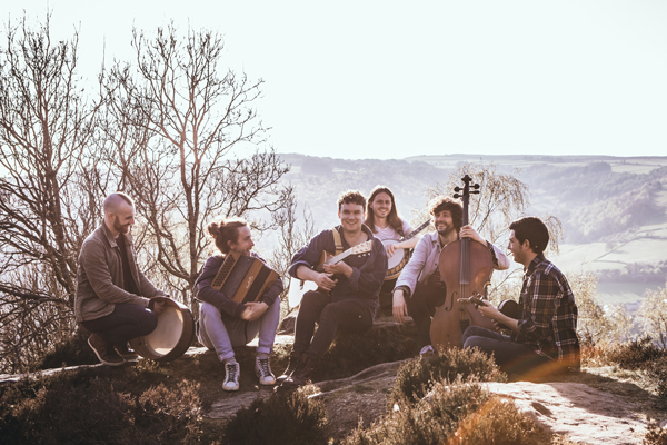 Sam Kelly & The Lost Boys image. The band are sitting down on top of a cliff. There are rolling green hills in the background. They are all sat together, smiling, with their instruments. 