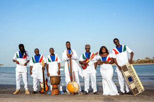 Suntou Susso & The Gambian Superstars image. The 7 members are stood on a sandy beach with the sea behind them. They have their instruments are are smiling.