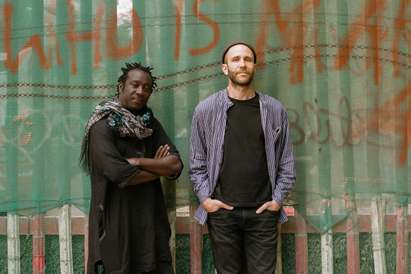 Touki image. The male duo are stood outside in front of a green wall that has red graffiti writing on it. 
