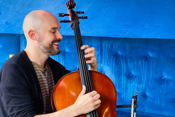 Arlo Anwin image. </p>
<p>We are looking at Arlo side on. He is playing the cello and smiling. He is a bald man with a short black beard. The background is bright blue (a bright blue sofa and bright blue painted wall). 
