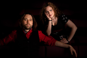 Antoine & Owena image. Antoine is sat on a sofa with his arms resting on the backrest. He is wearing a red shirt and a black waistcoat. Owena is sat behind the sofa on a chair. She is leaning forward with her head resting in her hand. She is wearing a black top, red trousers, and red lipstick. They are looking neutrally at the camera.