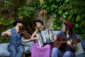 Clementine Lovell Trio image. The trio are sat outside with their instruments (accordion, fiddle, guitar).