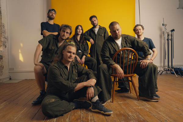 Danger Goat image. </p>
<p>The 7 piece band are mostly in sage green boilersuits, with a yellow backdrop. They are all looking casually directly into the camera. 
