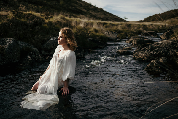 Detta Kenzie image. </p>
<p>Detta is in a flowy white dress in the countryside. She is sat leaning on a rock in the middle of a river. 