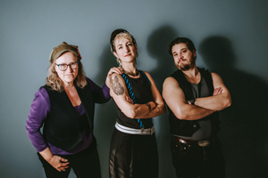 Dryadic image. The trio are stood in front of a grey / blue backdrop. They are lit well and we can see their shadows on the backdrop. They are wearing dark colours and have a punk / folkie style.