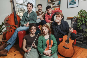 Evolution of Fishermen image. The 6 piece are sat in a living room. The 4 males are sat on the sofa and the females are sat in front of the sofa. They all have their instruments in the shot (double bass, flute, violin, guitar, banjo, drum)