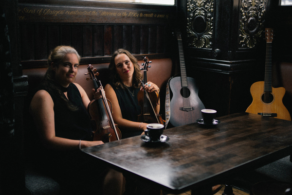 Grace Honeywell & Jeri Foreman image. </p>
<p>The duo are sat in a dimly lit pub holding their violins. Their guitars are in the background. They have gentle smiles. 