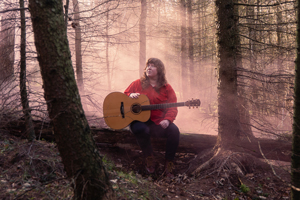 Holly Clarke Band image. Holly Clarke is sat in the foggy woods. There's a pink hue to the fog. She is holding her guitar and looking out into the distance.