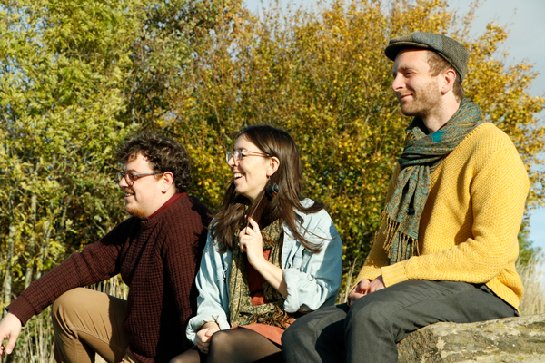 Lonan image. The trio are sat outside in the sunshine, smiling and looking into the distance. 