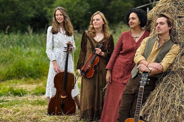 Meadowsuite image. </p>
<p>The 4 piece are stood in a field with their instruments. 2 of them are leaning about against a big stack of hay. 